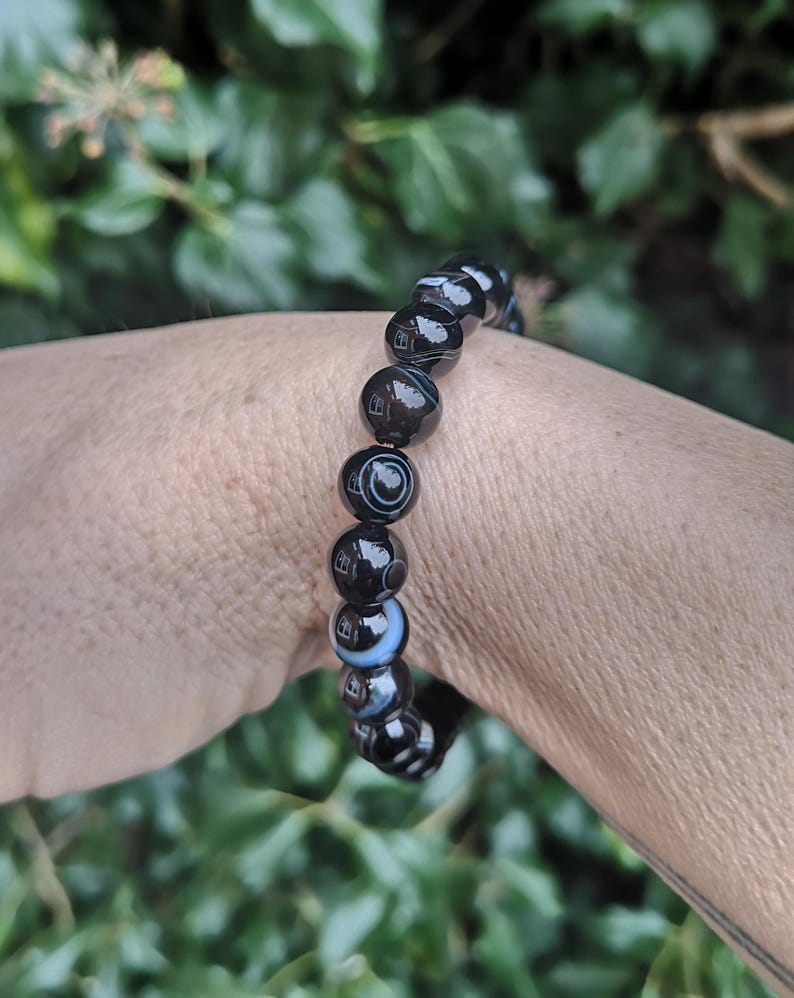 May include: A black and white beaded bracelet. The bracelet features round beads with a glossy finish. The beads have a marbled appearance with concentric circles and swirls. The bracelet is worn on a wrist.
