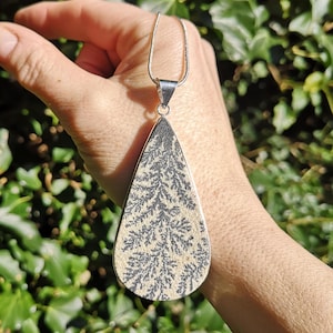 May include: A teardrop-shaped pendant necklace with a silver chain. The pendant features a cream-colored stone with a black, fern-like pattern, set in a silver bezel. The necklace is held against a backdrop of green foliage.