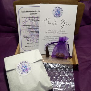 May include: An open cardboard box containing a white paper bag with a round sticker, a care card, a thank you card, a small purple bag, and a small wooden object. The cards have the words "Ascension Crystals" and "Thank You" printed on them. The background is purple.