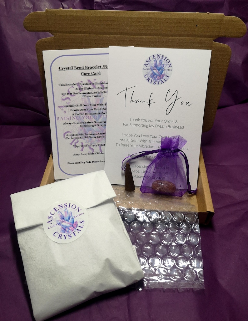 May include: An open cardboard box containing a white paper bag with a round sticker, a care card, a thank you card, a small purple bag, and a small wooden object. The cards have the words "Ascension Crystals" and "Thank You" printed on them. The background is purple.