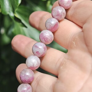 May include: A beaded bracelet featuring round, polished stones in varying shades of pink, magenta, and white. The beads are strung together in a single line, displaying a smooth, glossy texture with natural mineral inclusions throughout each stone.
