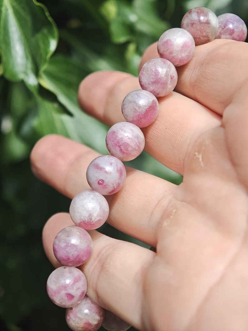 May include: A beaded bracelet featuring round, polished stones in varying shades of pink, magenta, and white. The beads are strung together in a single line, displaying a smooth, glossy texture with natural mineral inclusions throughout each stone.