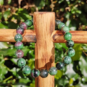 May include: A green and pink gemstone bracelet with round beads. The bracelet is displayed on a wooden cross.
