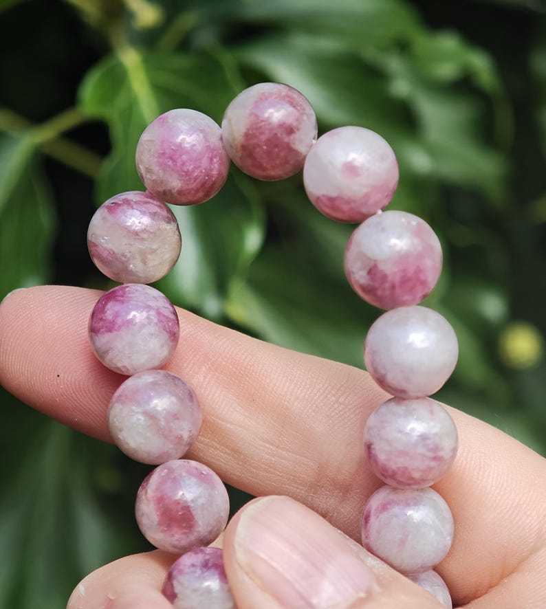 May include: A beaded bracelet featuring smooth, round gemstone beads in varying shades of pink, magenta, and white. Each bead displays a unique, marbled pattern with translucent and opaque sections, strung together to form a continuous circular loop.