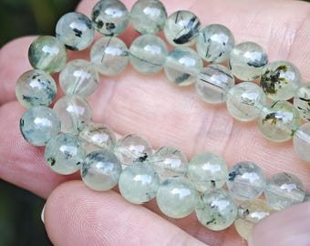 May include: A strand of polished, translucent light green beads featuring dark, needle-like inclusions throughout. The spherical beads are strung together, showcasing variations in clarity and the distinct black mineral patterns within each stone.