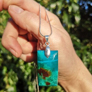 Pu&ograve; includere: Collana con pendente rettangolare in resina trasparente contenente una pietra turchese e marrone. Il pendente &egrave; appeso a una sottile catena d'argento. La pietra presenta un motivo naturale unico, lungo circa 3 cm.