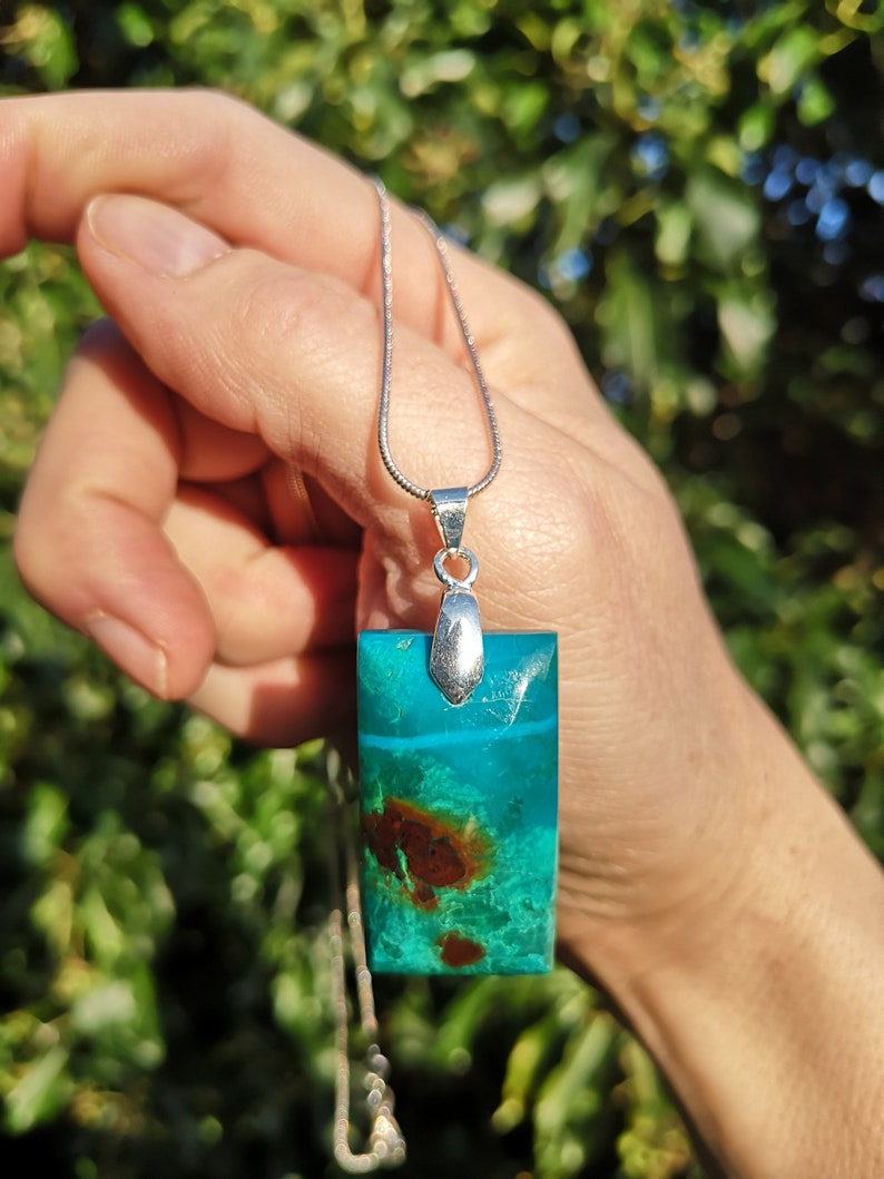 Pu&ograve; includere: Collana con pendente rettangolare in resina trasparente contenente una pietra turchese e marrone. Il pendente &egrave; appeso a una sottile catena d'argento. La pietra presenta un motivo naturale unico, lungo circa 3 cm.