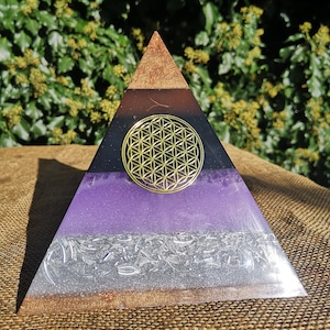 May include: A multi-layered pyramid made of resin with a flower of life design in gold on the black layer. The pyramid has layers of brown, black, purple, silver, and brown.
