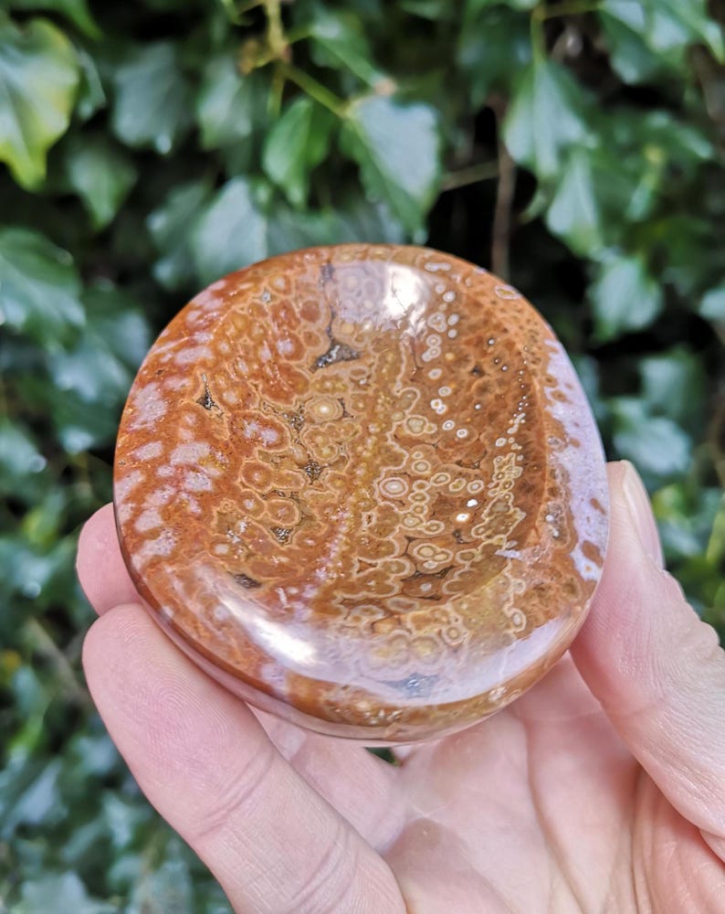 May include: A smooth, polished, oval-shaped, brown and white agate stone with a deep, bowl-like indentation. The stone has a swirling pattern of white and brown.
