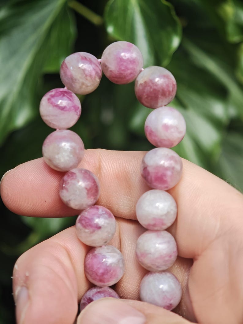 May include: A strand of round, polished beads featuring a marbled pattern of vibrant magenta, soft pink, and opaque white. The beads are strung together in a vertical arrangement, showcasing a smooth, glossy texture and varied color distribution.