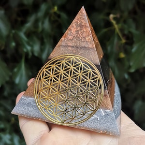 May include: A pyramid-shaped resin ornament with a gold flower of life design on a black background. The pyramid is layered with brown, copper, and silver glitter.