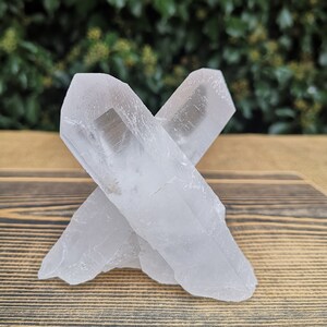 May include: Two clear quartz crystals, one lying on top of the other, forming an X shape. The crystals are on a wooden surface.