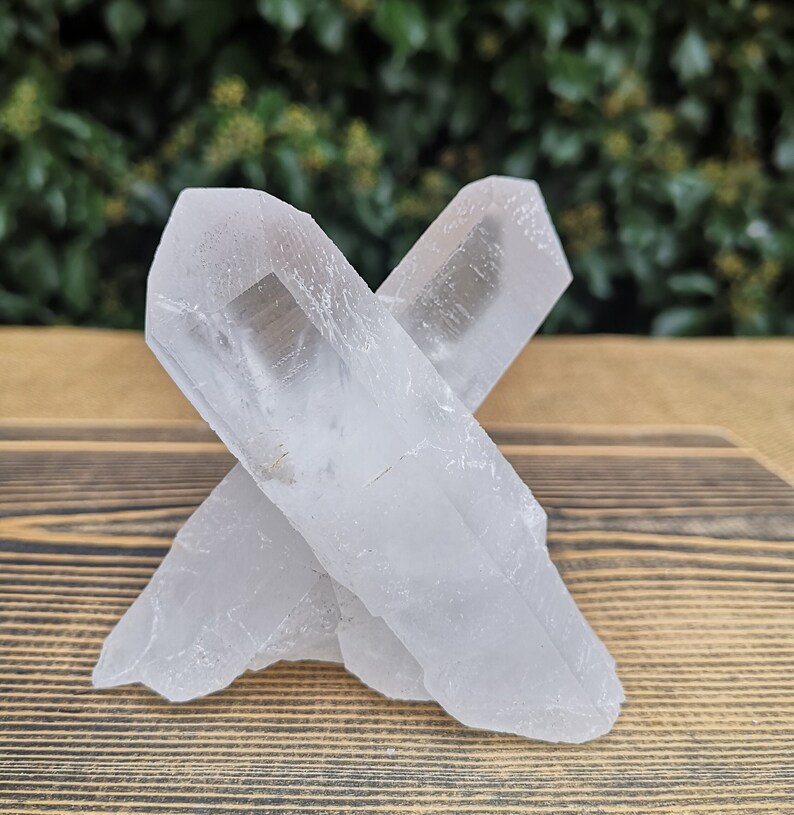 May include: Two clear quartz crystals, one lying on top of the other, forming an X shape. The crystals are on a wooden surface.