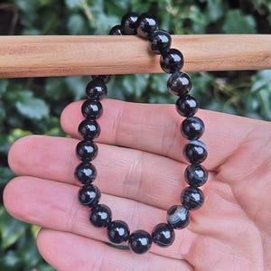 May include: A black beaded bracelet with a glossy finish. The bracelet is made of round, polished beads, strung together. The beads have a marbled appearance with subtle gray and white patterns. The bracelet is held against a blurred green background.