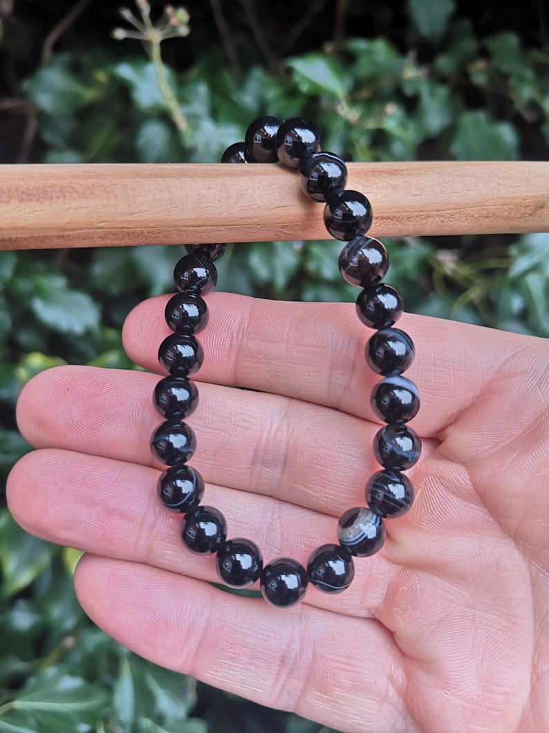 May include: A black beaded bracelet with a glossy finish. The bracelet is made of round, polished beads, strung together. The beads have a marbled appearance with subtle gray and white patterns. The bracelet is held against a blurred green background.