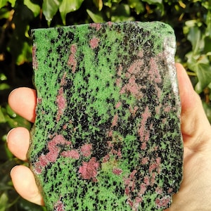 May include: A hand displays a rough-cut stone. The stone has a green base, with black and pinkish-red speckles. The surface is uneven, highlighting its natural texture. The background is blurred, with green foliage.