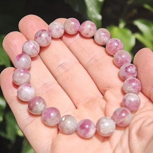May include: A stretch bracelet composed of round, polished beads in varying shades of pink, magenta, and white. The beads feature natural marbling and translucent inclusions, creating a unique pattern on each stone throughout the circular design.