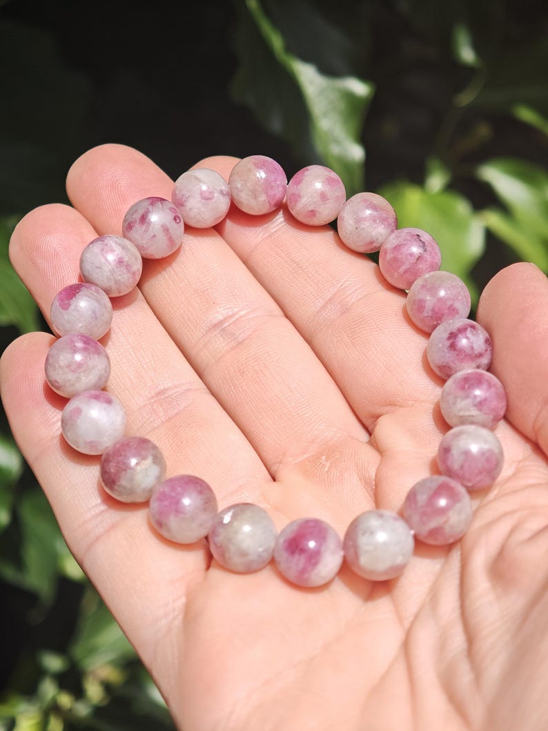 May include: A stretch bracelet composed of round, polished beads in varying shades of pink, magenta, and white. The beads feature natural marbling and translucent inclusions, creating a unique pattern on each stone throughout the circular design.