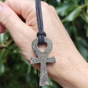 May include: A metallic ankh pendant with a textured, dark grey, pyrite-like finish, suspended from a simple black cord. The cross-shaped charm features a circular loop at the top and a vertical base, showcasing a natural, crystalline surface texture.