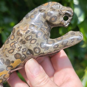 May include: A stone leopard figurine with a spotted pattern in brown, tan, and grey. The sculpture is held in a hand, with green foliage in the background. The leopard is carved with detail.