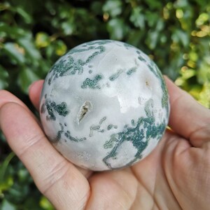 May include: A green and white agate sphere, polished to a smooth finish. The sphere has a swirling pattern of green and white, with a few small white spots.