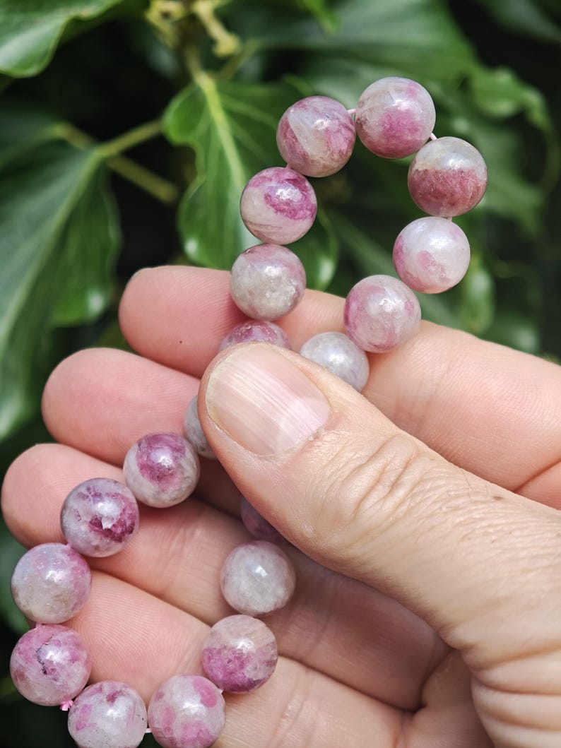 May include: A strand of round, polished beads featuring a mix of translucent white and vibrant pink or magenta hues. The beads are strung together in a continuous line, showcasing natural variations in color and internal texture within each stone.
