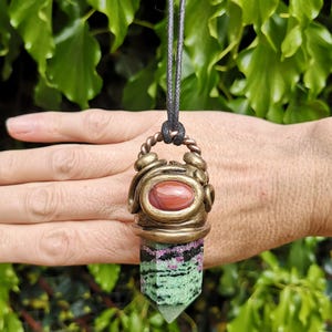 May include: A pendant necklace featuring a gemstone pendant with a red oval stone and a green and black crystal. The pendant is suspended from a black cord. The metalwork has a bronze finish.