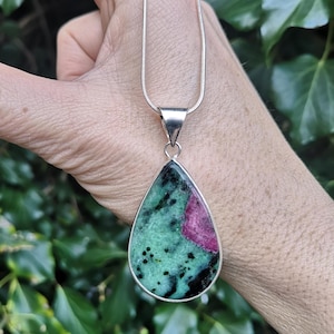 May include: A silver necklace with a teardrop-shaped pendant. The pendant has a green and black stone with a splash of pink. The necklace is set against a backdrop of green foliage.