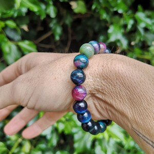 May include: A bracelet made of round, multi-colored beads. The beads are a mix of blue, green, and pink with a shimmery finish. The bracelet is on a person's wrist.