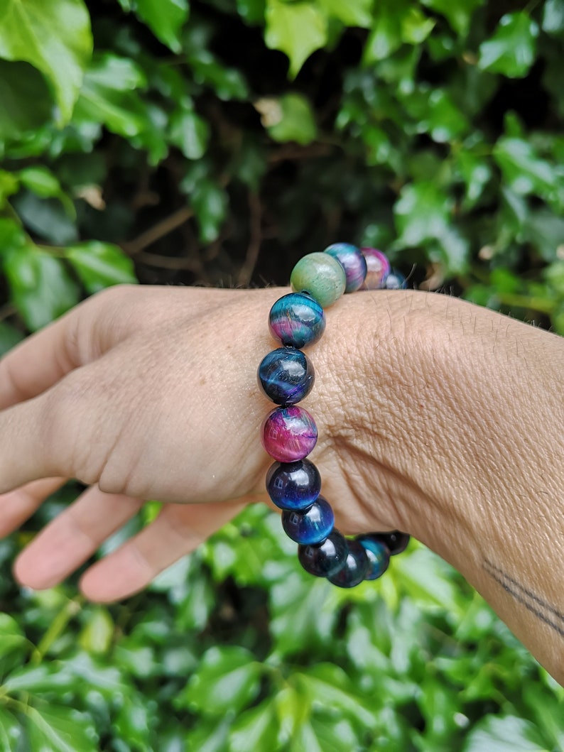 May include: A bracelet made of round, multi-colored beads. The beads are a mix of blue, green, and pink with a shimmery finish. The bracelet is on a person's wrist.