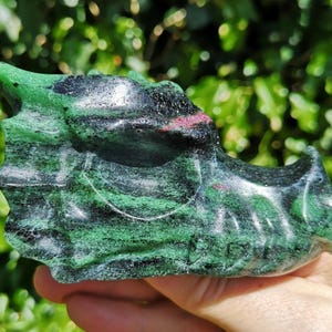 May include: A carved dragon head sculpture made of green and black stone with red accents. The sculpture is held in a hand and has a detailed, textured surface. The dragon's head is facing left, with its mouth slightly open.