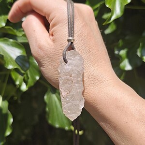 May include: A clear, raw, jagged crystal pendant suspended from a dark grey cord. The crystal has an irregular, multifaceted surface texture and a pointed base. A small, smooth, silver-toned metal bead secures the cord above the crystal stone.