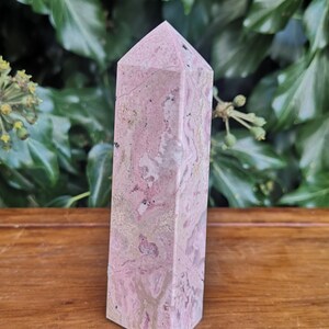 May include: A pink and white polished crystal point, also known as a crystal wand, with a natural, rough texture. The crystal is on a wooden surface.