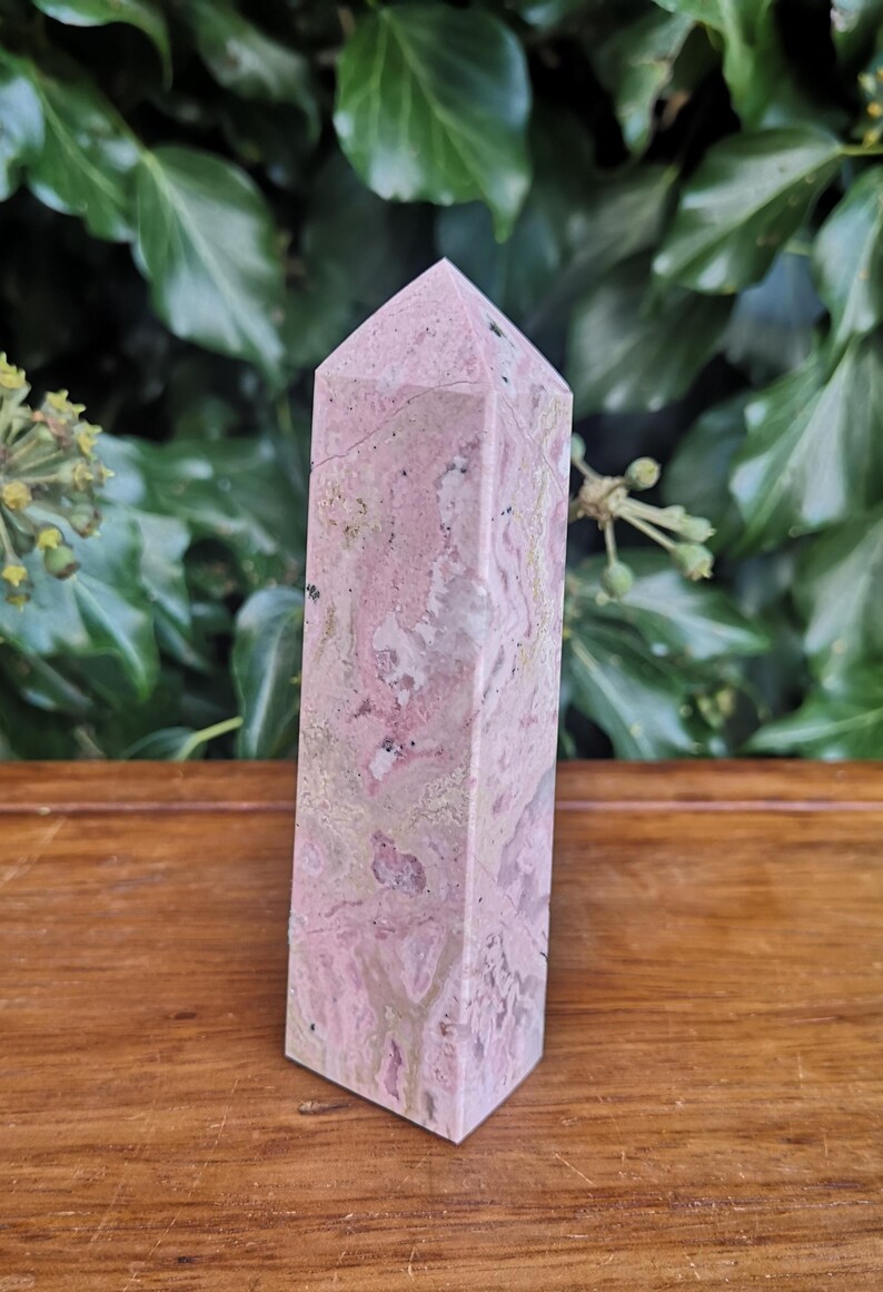 May include: A pink and white polished crystal point, also known as a crystal wand, with a natural, rough texture. The crystal is on a wooden surface.