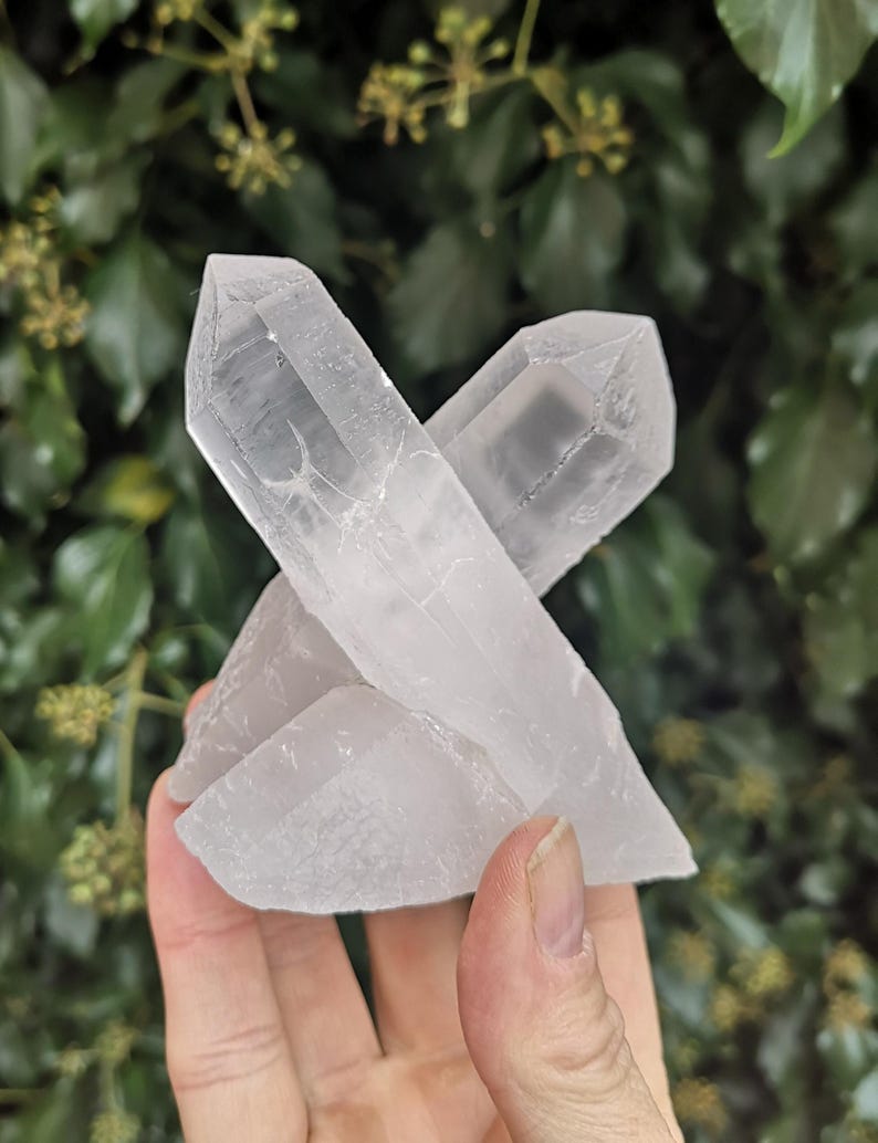 May include: A cluster of three clear quartz crystals with a pointed shape. The crystals are arranged in a way that they appear to be stacked on top of each other.