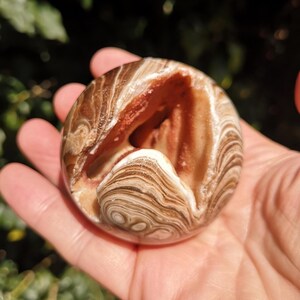 K&ouml;nnte beinhalten: Eine braune und wei&szlig;e Achatkugel mit einer nat&uuml;rlichen &Ouml;ffnung, die ein geschichtetes Inneres enth&uuml;llt. Die Kugel ist glatt und poliert, mit einem wirbelnden Muster aus Braun, Wei&szlig; und Orange.