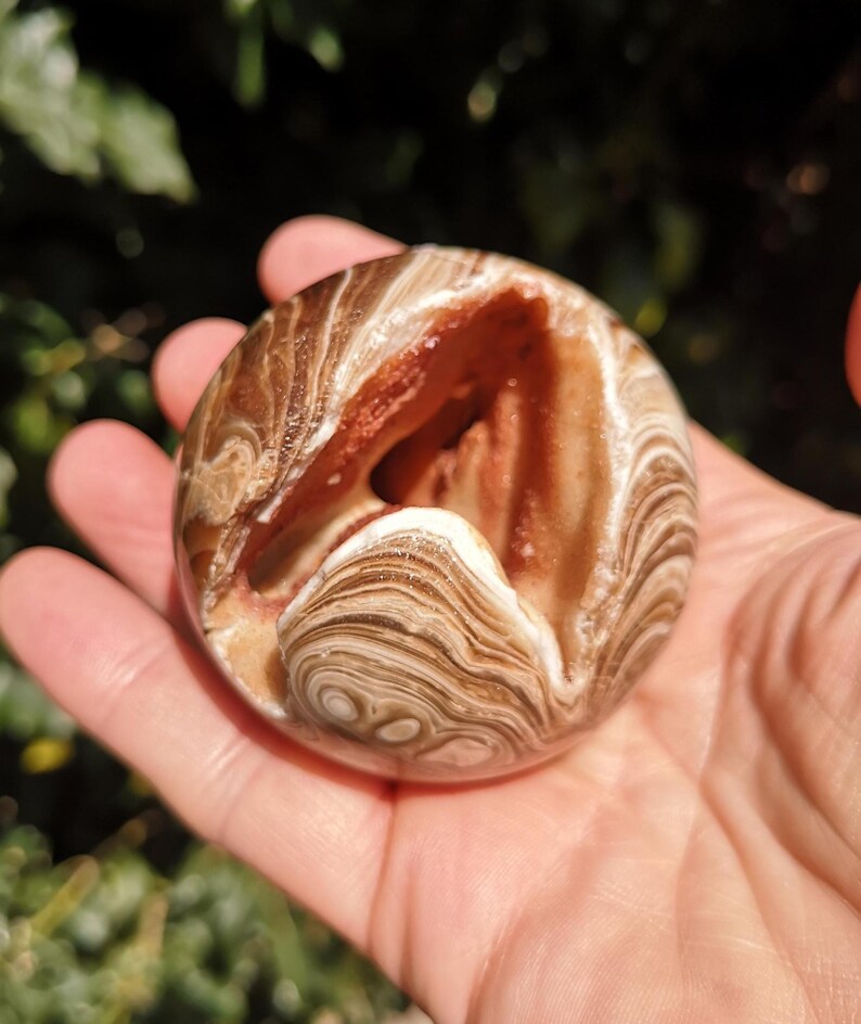 K&ouml;nnte beinhalten: Eine braune und wei&szlig;e Achatkugel mit einer nat&uuml;rlichen &Ouml;ffnung, die ein geschichtetes Inneres enth&uuml;llt. Die Kugel ist glatt und poliert, mit einem wirbelnden Muster aus Braun, Wei&szlig; und Orange.