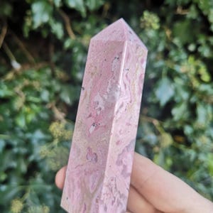 May include: A pink and white polished crystal point, also known as a wand, made of rhodochrosite. The crystal has a natural, marbled pattern.