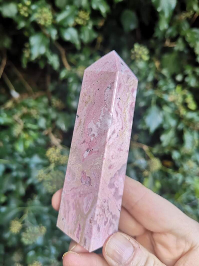 May include: A pink and white polished crystal point, also known as a wand, made of rhodochrosite. The crystal has a natural, marbled pattern.