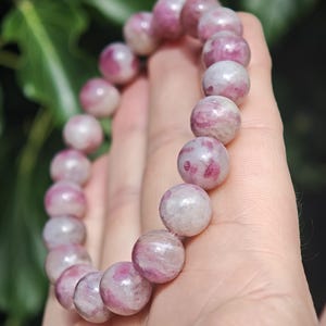 May include: A beaded bracelet composed of smooth, spherical stones featuring a marbled pattern of opaque white and vibrant pink hues. The beads are strung together in a circular formation, displaying natural variations in color and texture throughout.