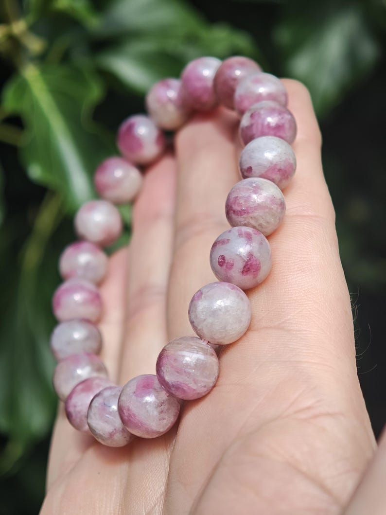 May include: A beaded bracelet composed of smooth, spherical stones featuring a marbled pattern of opaque white and vibrant pink hues. The beads are strung together in a circular formation, displaying natural variations in color and texture throughout.