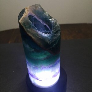 May include: A green, purple, and blue fluorite crystal lamp with a white light shining through the crystal. The lamp is on a black base.