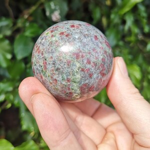 May include: A polished, spherical gemstone with a speckled pattern of red and green hues. The stone is held in a hand, showcasing its smooth surface and natural colours. This decorative sphere is a unique addition to any collection.