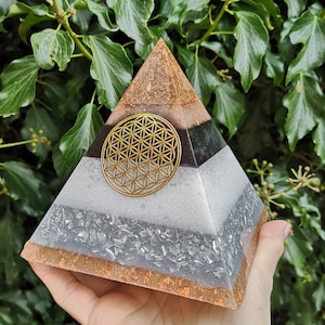 May include: A multi-layered pyramid made of clear resin with gold leaf accents. The pyramid has a black layer with a gold flower of life symbol in the center. The pyramid is a popular item for home decor and spiritual practices.