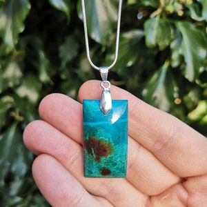 Pu&ograve; includere: Una collana con pendente rettangolare turchese con inclusioni marroni e verdi, sospesa a una catena d'argento. Il pendente &egrave; tenuto in mano su uno sfondo di fogliame verde. Un gioiello elegante.