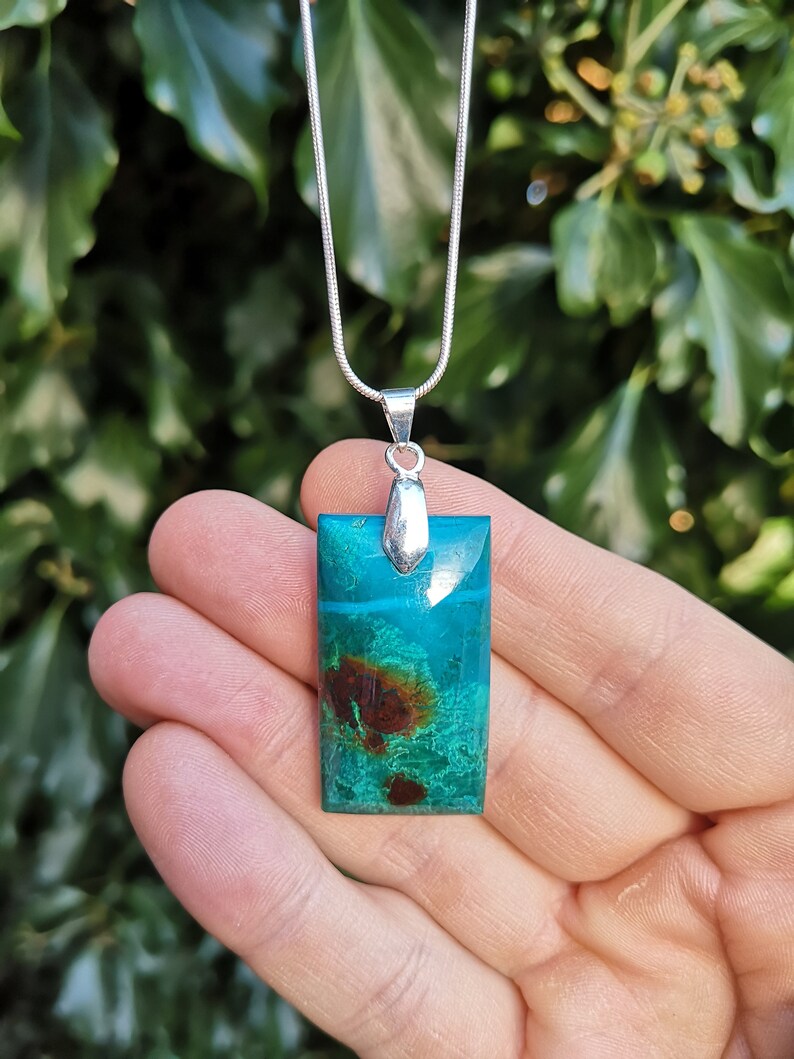 Pu&ograve; includere: Una collana con pendente rettangolare turchese con inclusioni marroni e verdi, sospesa a una catena d'argento. Il pendente &egrave; tenuto in mano su uno sfondo di fogliame verde. Un gioiello elegante.