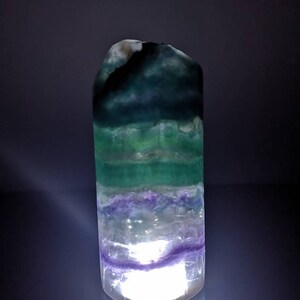 May include: A green, purple, and white fluorite crystal with a light shining through it. The crystal is shaped like a tower and has a smooth, polished surface.