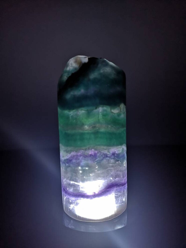 May include: A green, purple, and white fluorite crystal with a light shining through it. The crystal is shaped like a tower and has a smooth, polished surface.