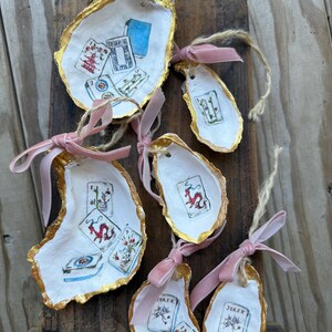 May include: Hand-painted oyster shell ornaments with playing card designs. Each shell is edged in gold and adorned with a pink or gray ribbon for hanging. The ornaments feature various card suits and a joker.
