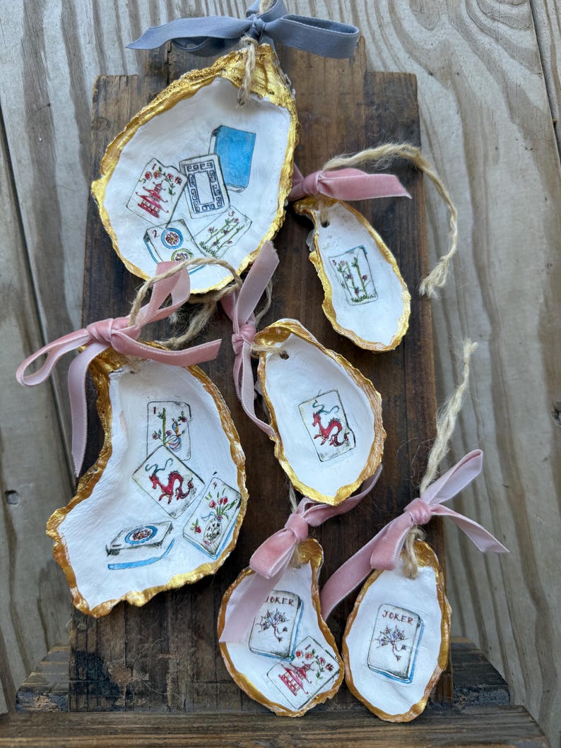 May include: Hand-painted oyster shell ornaments with playing card designs. Each shell is edged in gold and adorned with a pink or gray ribbon for hanging. The ornaments feature various card suits and a joker.